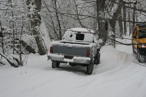 1990 Chevrolet Suburban - A gray pickup truck is parked in a snowy forest, with a yellow SUV partially visible on the right.