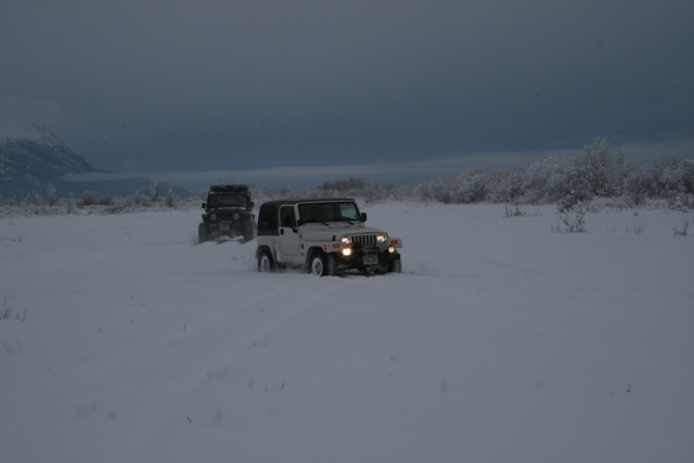 White Jeep Wrangler driving in snow, dark Jeep behind it