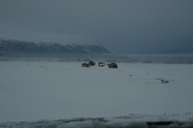 Several vehicles drive across a vast, snow-covered plain with mountains in the background under a cloudy sky
