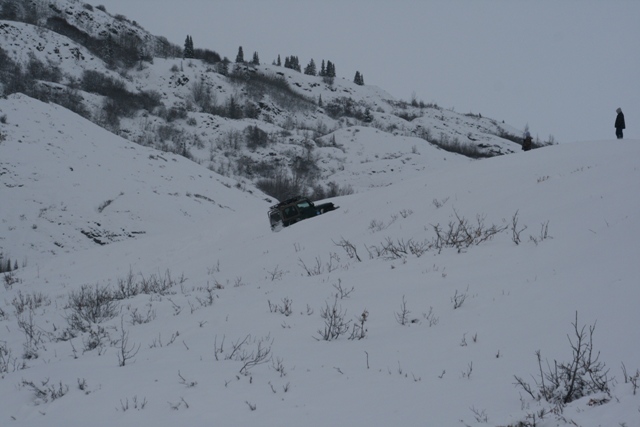 Dark SUV navigating a snow-covered hillside with a person standing on a ridge above