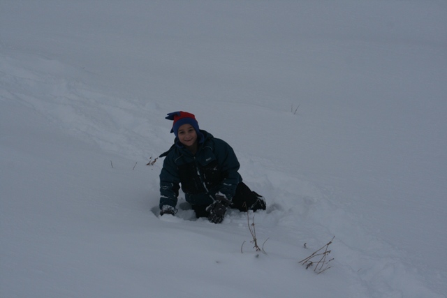 Kid in a blue and red hat and a dark jacket is crawling in deep snow