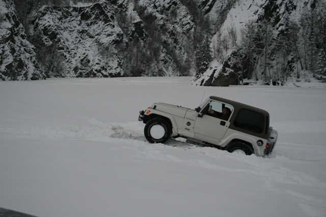 White Jeep Wrangler driving through deep snow near a rocky, snow-covered mountainside