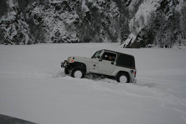 White Jeep Wrangler with brown hardtop driving through deep snow near a rocky, snow-covered hillside