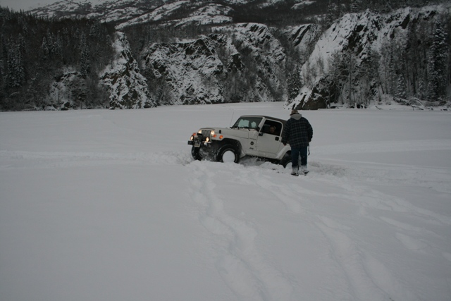 White Jeep Wrangler stuck in deep snow, driver standing beside it. Mountains and trees in background