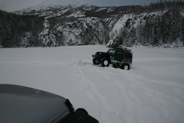 Dark green Jeep Wrangler with roof rack driving in deep snow, another vehicle's hood visible in foreground