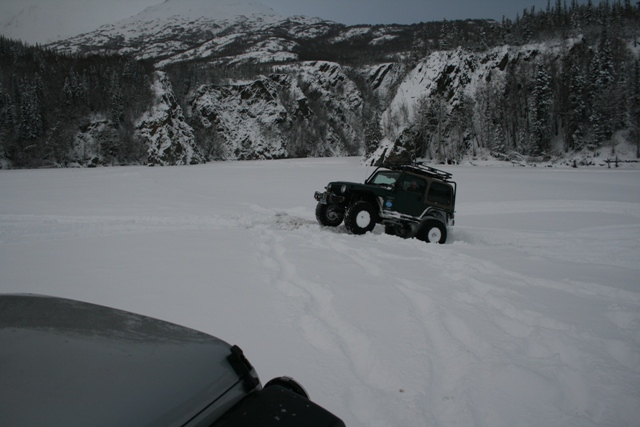 Dark green Jeep Wrangler with roof rack driving in snow, with another vehicle in the foreground