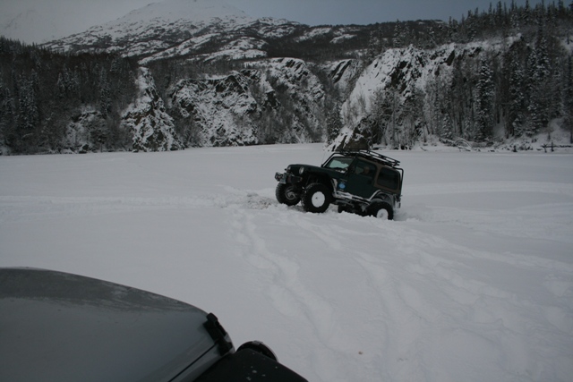 Dark green Jeep Wrangler driving through deep snow with mountains in background