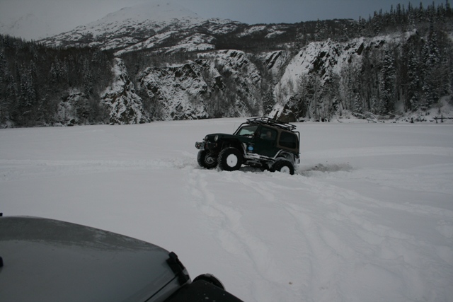 Jeep Wrangler driving through deep snow with a mountain and forest in the background. Another vehicle is partially visible in the foreground