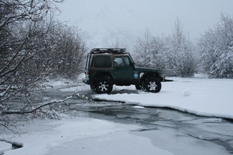 1990 Chevrolet Suburban - A green Jeep Wrangler navigates through a partially frozen stream surrounded by snow-covered trees and brush.
