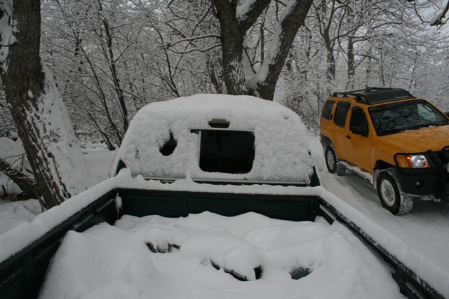 Snow covers the bed of a dark green pickup truck. A yellow SUV is parked to the right on a snow-covered road