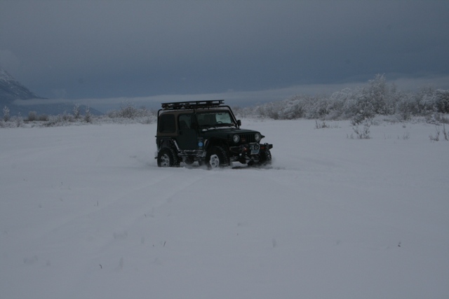 Dark green Jeep Wrangler with roof rack driving through deep snow, trees and mountains in background