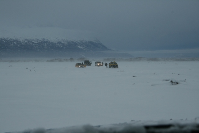 Several SUVs with headlights on drive across a snow-covered plain towards a misty mountain range