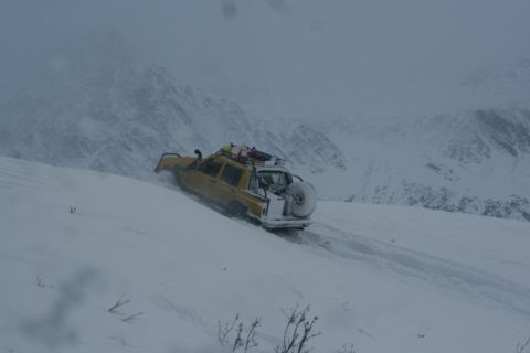 1990 Chevrolet Suburban - A yellow SUV navigates a steep, snow-covered mountain slope, its roof rack laden with gear and a spare tire mounted at the rear.