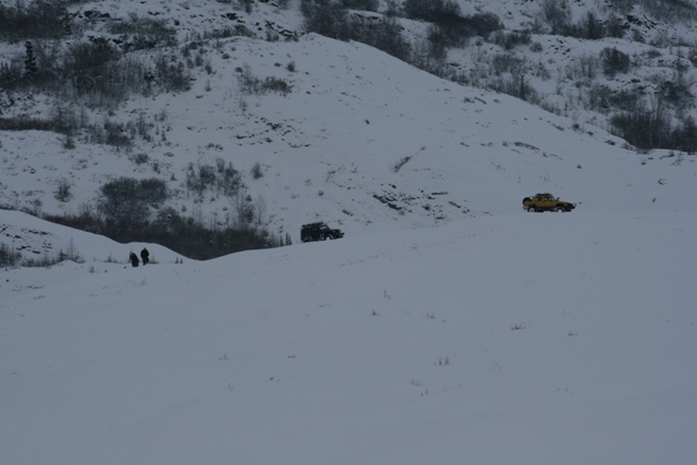 Two vehicles, one dark and one yellow, drive across a snow-covered hill with people standing nearby