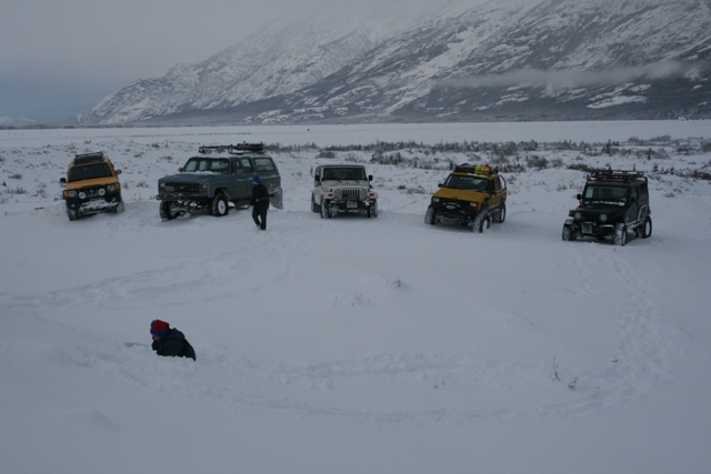 Five off-road vehicles parked in deep snow with mountains in the background. One person is walking between two vehicles, another person is partially buried in snow