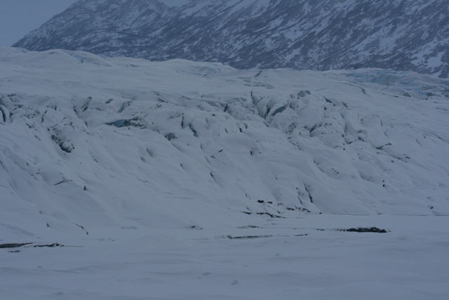 Snow-covered glacier with crevasses, leading to snow-dusted mountains in the background