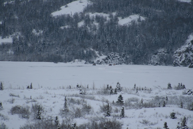 Two vehicles on a frozen, snow-covered lake with forested hills in the background