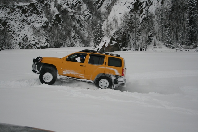 Yellow SUV with roof rack driving through snow, kicking up powder