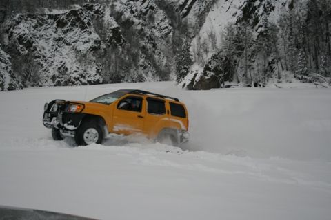 1990 Chevrolet Suburban - A yellow SUV navigates a snow-covered landscape, with steep, tree-lined mountains visible behind it.