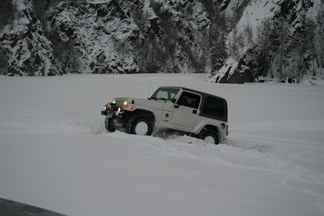 White Jeep Wrangler driving through deep snow near a rocky, tree-covered mountainside