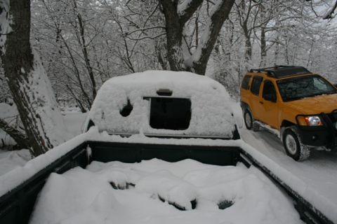 1990 Chevrolet Suburban - A snow-covered truck bed is in the foreground with a yellow SUV driving on a snowy road in the background.