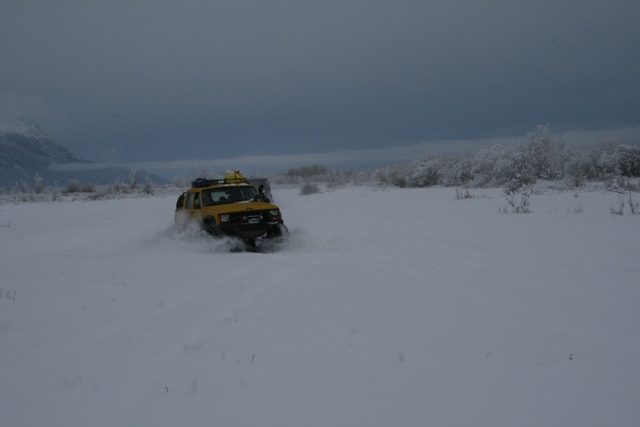 Yellow SUV with roof rack driving through deep snow, kicking up spray