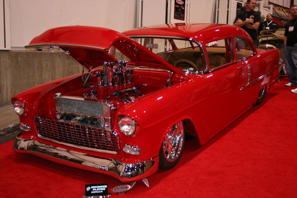 Red 1955 Chevrolet Bel Air hot rod with hood open, showing a large engine and chrome details, on a red carpet