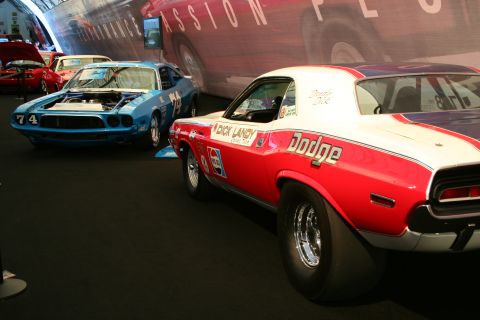 Rear view of a red and white Dodge Challenger drag car with wide rear tires. A blue Dodge Challenger with numb