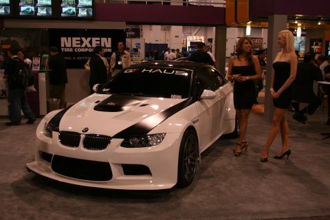 White BMW M3 coupe with black hood and roof stripes, two women standing nearby at an indoor event
