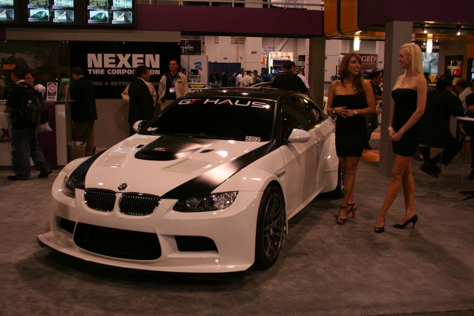 White BMW M3 coupe with black hood and roof stripes, two women standing nearby at an indoor event