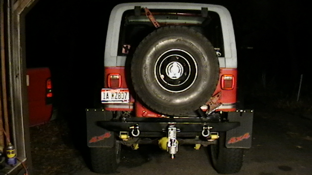 Rear view of a red and white Jeep with a spare tire on the back, aftermarket bumper, and mud flaps. License plate reads 1A WZ807