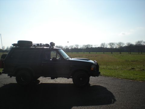 1993 Ford Bronco - A dark-colored Ford Bronco is parked on a road next to a grassy field under a bright sky.