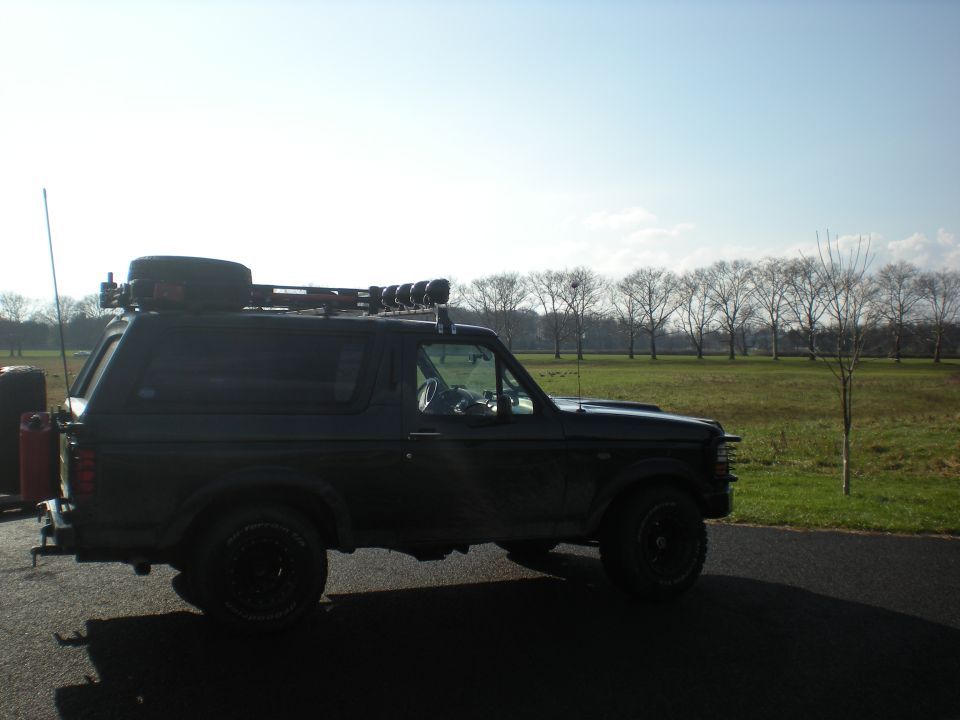 Black Ford Bronco with roof rack, spare tire, and auxiliary lights parked on asphalt
