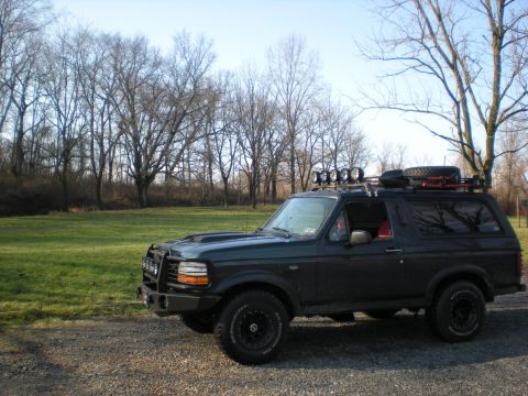 1993 Ford Bronco - A modified Ford Bronco sits parked on a gravel surface with a grassy field and bare trees in the background.
