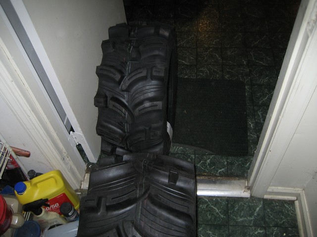 Stack of aggressive lugged ATV mud tires leaned against a wall next to oil jugs