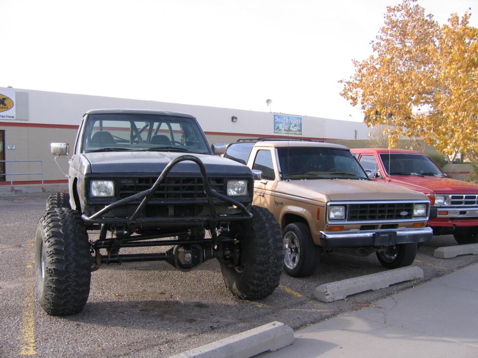 Front view of a lifted, dark-colored truck with a roll cage and bull bar, parked next to a tan SUV and a red SUV