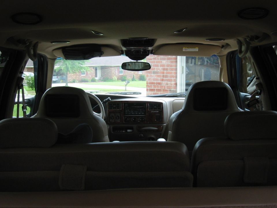 View from rear of SUV interior showing tan seats, headrest video screens, dash, and windshield looking out at a house