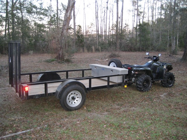Green ATV towing a utility trailer with wood deck and toolbox in a wooded area