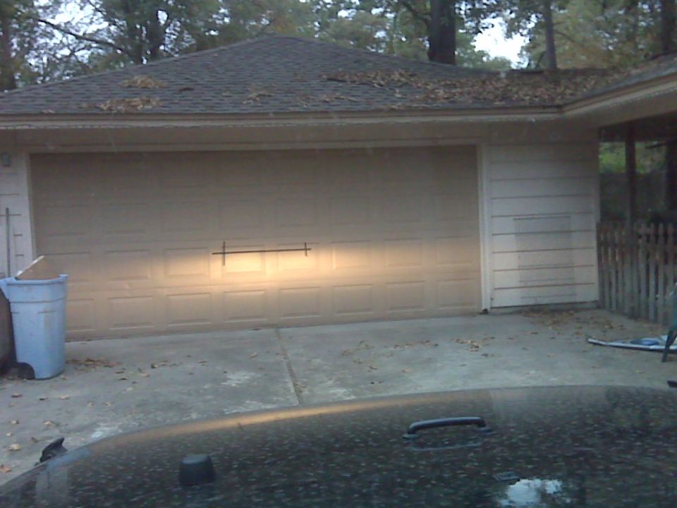 Car hood in foreground, facing a garage door with headlights on. Trash can to the left
