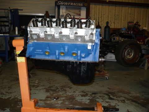 1973 Ford Ranchero - V8 engine on an engine stand in a workshop, with a partial vehicle chassis in the background.
