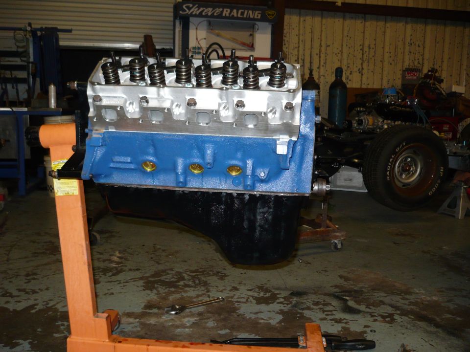 V8 engine block painted blue, with aluminum heads and valve springs, mounted on an orange engine stand. Another vehicle chassis is visible in the background
