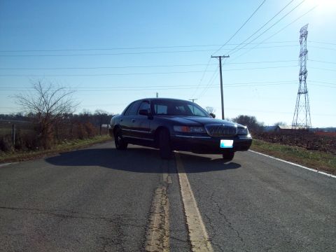 1999 Mercury Grand Marquis - A dark sedan is parked on a two-lane road with a power line tower in the background.
