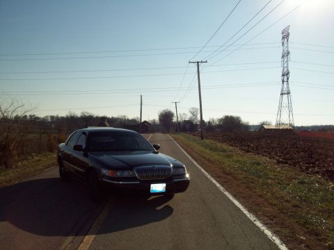 1999 Mercury Grand Marquis - A dark sedan is parked on the side of a road with fields and power lines in the background.