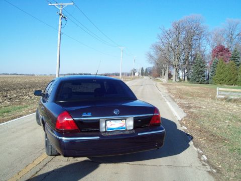 1999 Mercury Grand Marquis - A dark blue sedan is parked on the side of a rural road under a clear blue sky.