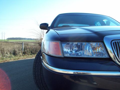 1999 Mercury Grand Marquis - The front corner of a dark sedan is shown, with a field and power lines in the background. The website 'SuperMotors.net' is visible at the…
