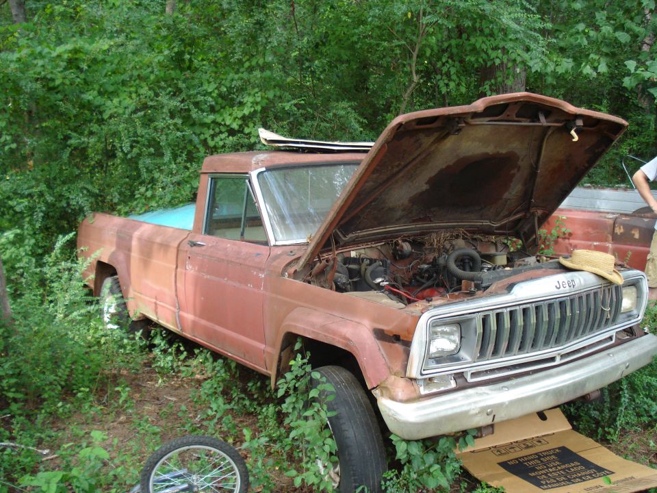 Rusty red Jeep J-10 pickup truck with hood open, parked in overgrown brush. A straw hat rests on the engine
