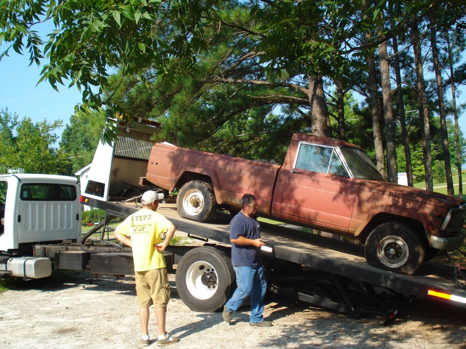 Brown Jeep J-10 pickup truck on a flatbed tow truck, with a white tow truck in the background. Two men are standing nearby