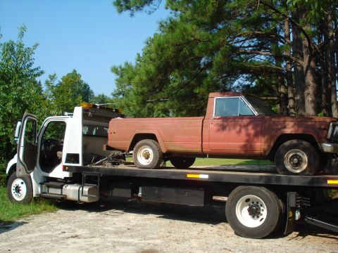 1984 Jeep J-10 - A rusty Jeep pickup truck is being transported on a flatbed tow truck under a clear blue sky.