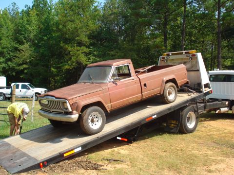 1984 Jeep J-10 - A weathered brown pickup truck is winched onto a tow truck, with another white truck visible in the background.