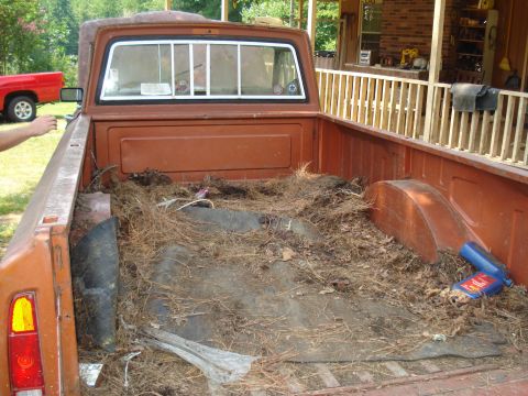 1984 Jeep J-10 - The bed of an orange 1984 Jeep J-10 pickup truck is filled with pine needles and debris, with another red truck visible in the background.